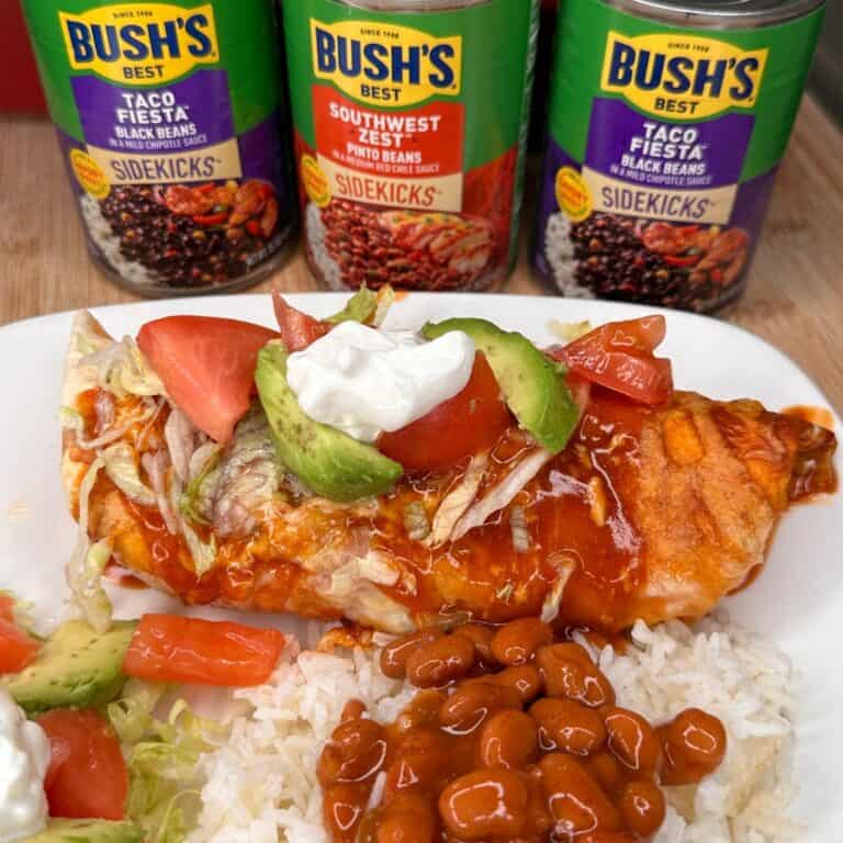 A plate with an enchilada topped with avocado, tomato, lettuce, sour cream, and sauce, served with rice and beans. Three cans of Bush’s Best beans are in the background.
