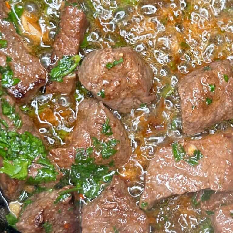Close-up of sizzling beef cubes cooking in a pan, garnished with chopped parsley and surrounded by bubbling oil and seasonings.