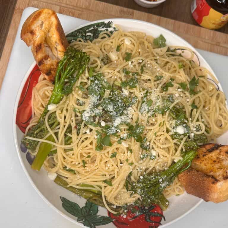 A plate of spaghetti with broccolini, garnished with grated cheese and herbs, served with two pieces of toasted bread on the side. The dish is on a colorful plate and placed on a wooden surface.