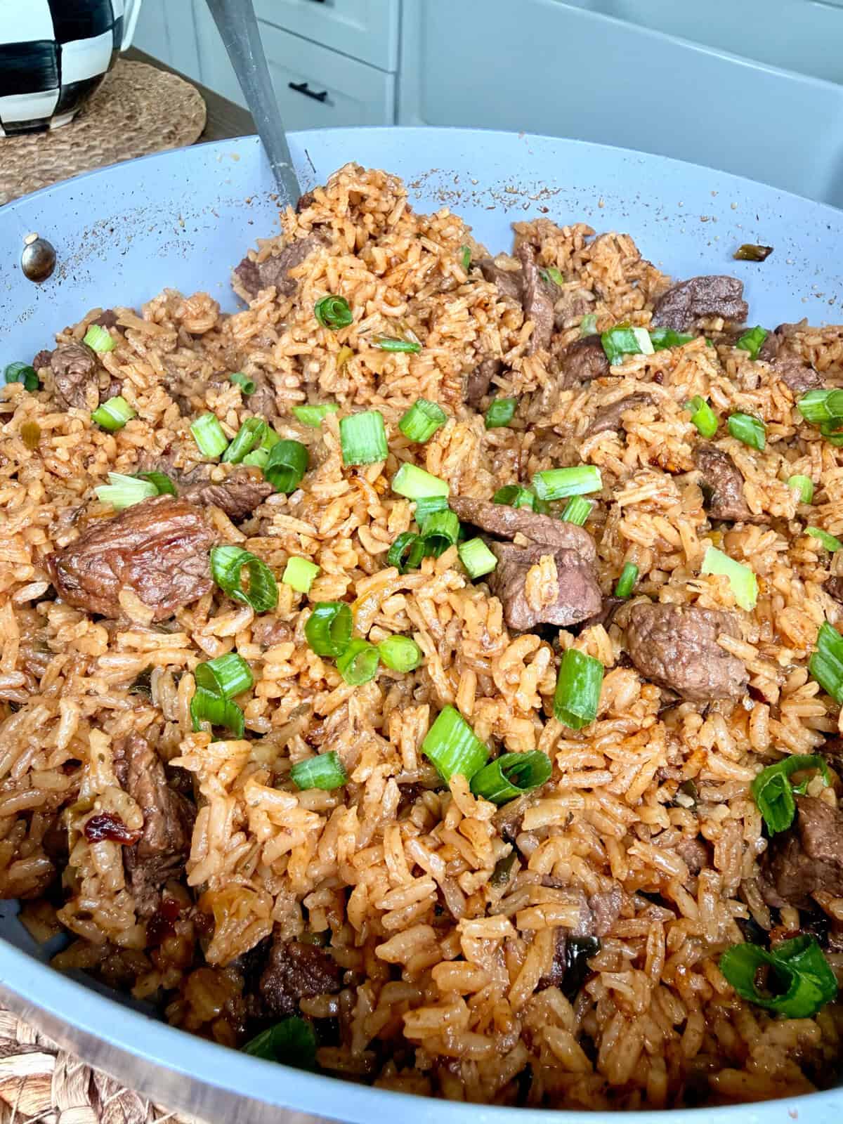 A close-up of the Best One Pot Steak and Rice shows a skillet brimming with seasoned rice, juicy beef chunks, and chopped green onions, all freshly cooked and garnished, with a serving spoon resting in the pan.
