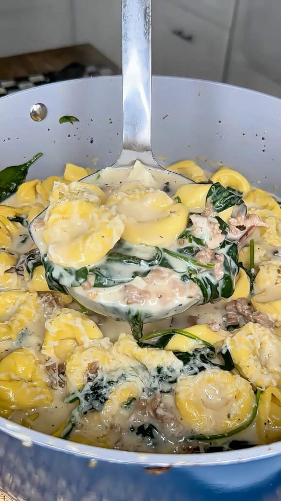 A ladle scoops creamy cheese-filled tortellini with spinach and ground meat from a pan, showing a rich, cheesy sauce coating the pasta.
