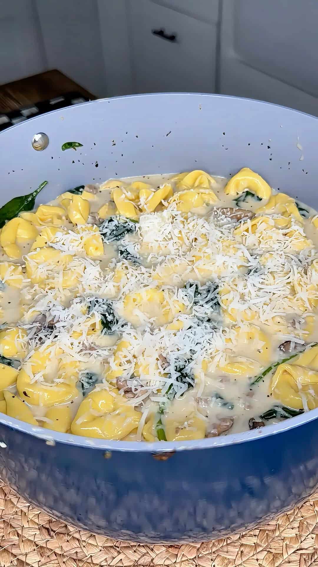 A skillet filled with cheese tortellini in a creamy sauce, mixed with spinach and mushrooms, and topped with freshly grated parmesan cheese.
