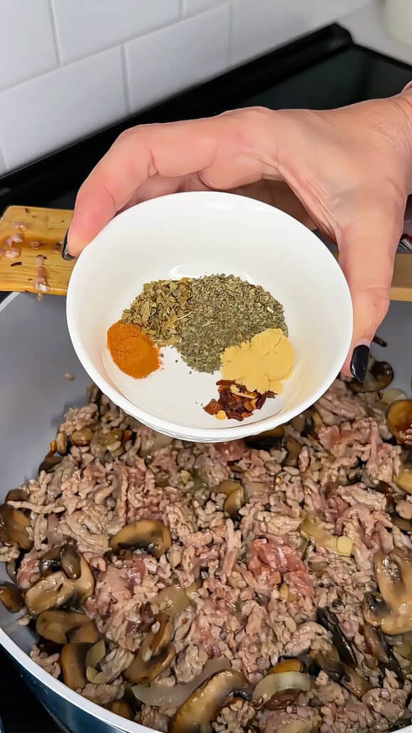 A hand holds a white bowl with four different seasonings over a pan of ground meat and mushrooms cooking on a stovetop.