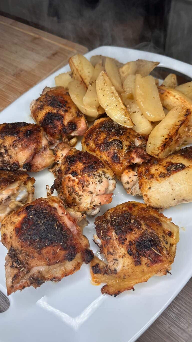 A white plate with several pieces of roasted, seasoned chicken and golden-brown potato wedges, set on a wooden surface. Steam rises from the food, bringing to mind the warmth of a freshly made pizza recipe straight from the oven.