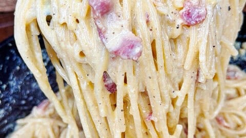 A close-up of creamy spaghetti carbonara being lifted with a spoon, showing strands of pasta coated in sauce with pieces of bacon or pancetta, in a kitchen setting.