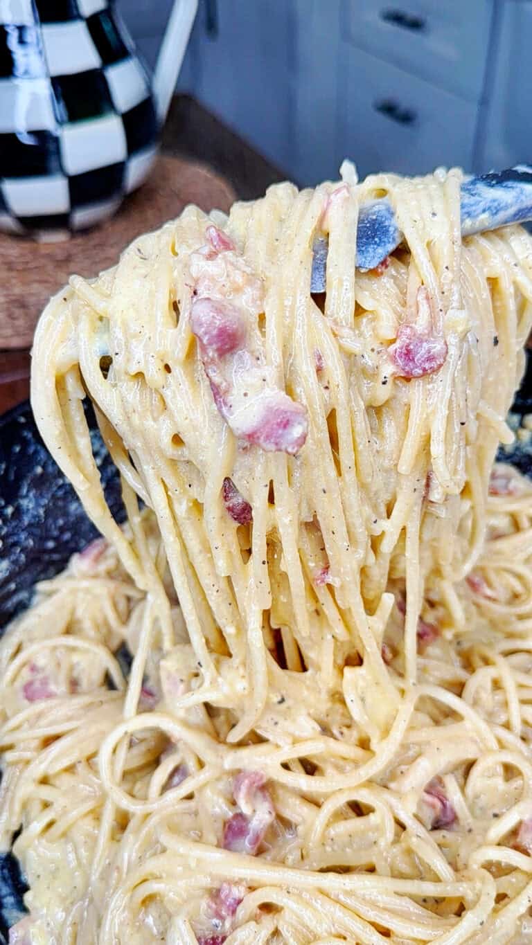 A close-up of creamy spaghetti carbonara being lifted with a spoon, showing strands of pasta coated in sauce with pieces of bacon or pancetta, in a kitchen setting.