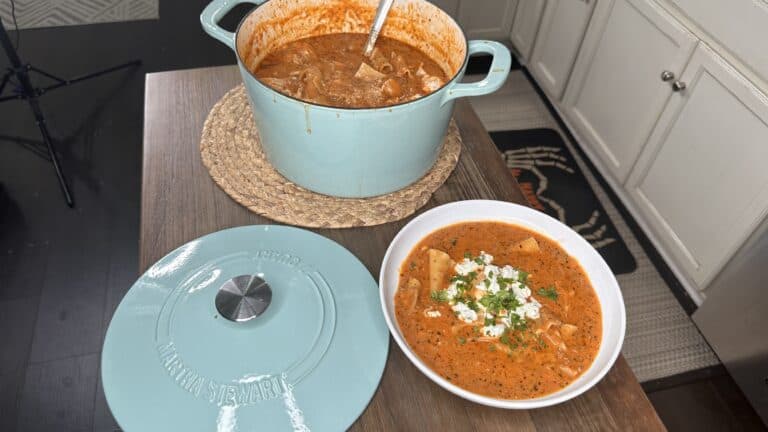 A blue Dutch oven filled with Lasagna Soup sits on a mat next to its lid and a white bowl of the same soup, topped with cheese and chopped herbs, on a wooden countertop in a kitchen.