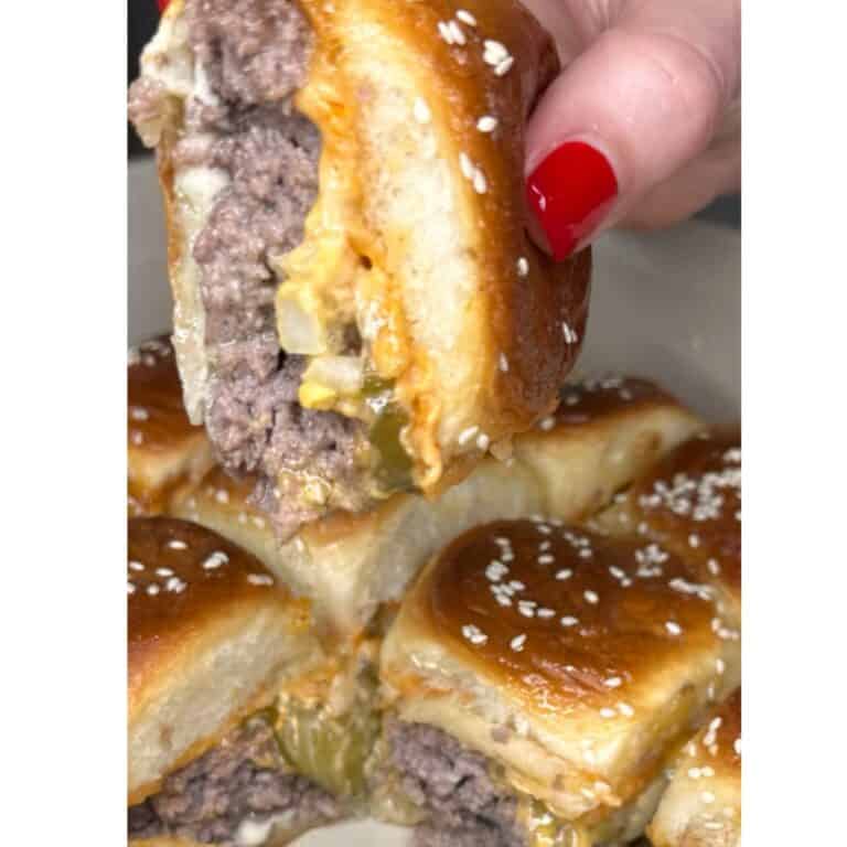 A close-up of a hand with red nail polish holding a cheesy slider made with Copy Cat Big Mac Sauce, a sesame seed bun, ground beef patty, onions, and pickles, with several more sliders stacked below.
