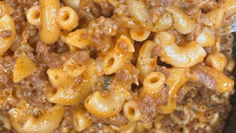 A pot filled with cooked macaroni, ground beef, and a rich tomato-based sauce—reminiscent of classic Hamburger Helper—rests on a woven mat. A metal spoon is partially visible, stirring the hearty mixture.