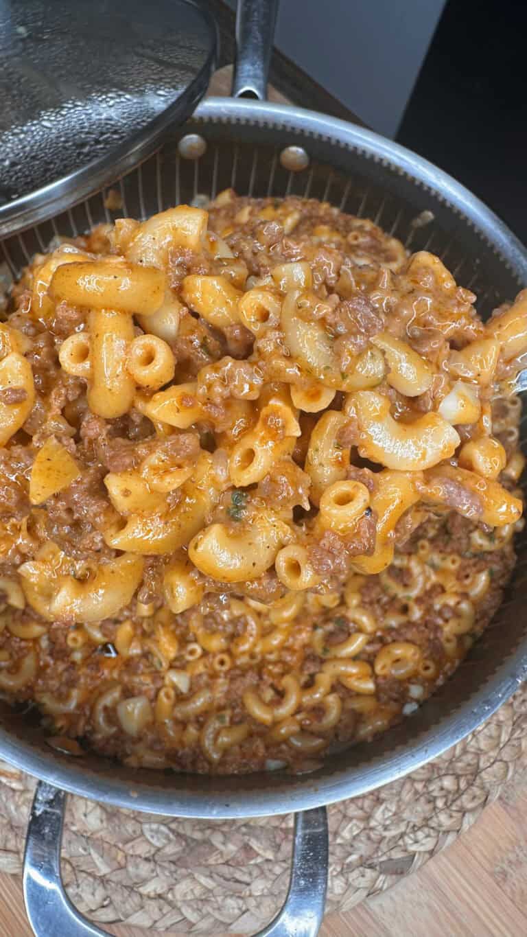 A pot filled with cooked macaroni, ground beef, and a rich tomato-based sauce—reminiscent of classic Hamburger Helper—rests on a woven mat. A metal spoon is partially visible, stirring the hearty mixture.