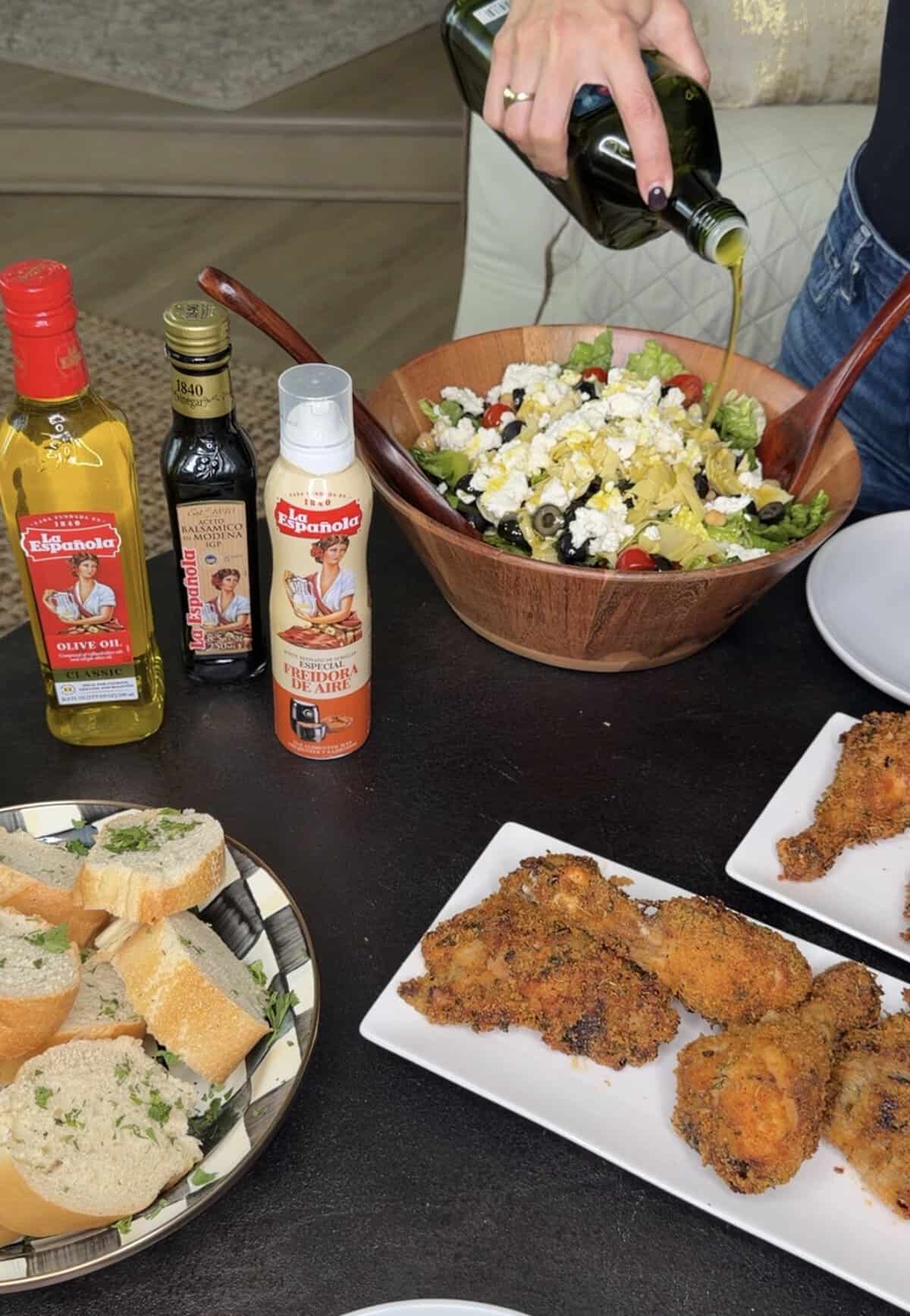 A hand pours olive oil on a salad in a wooden bowl. Around it are bottles of olive oil, balsamic glaze, and cooking spray, plus plates of sliced bread and crispy Italian Fried Chicken drumsticks.
