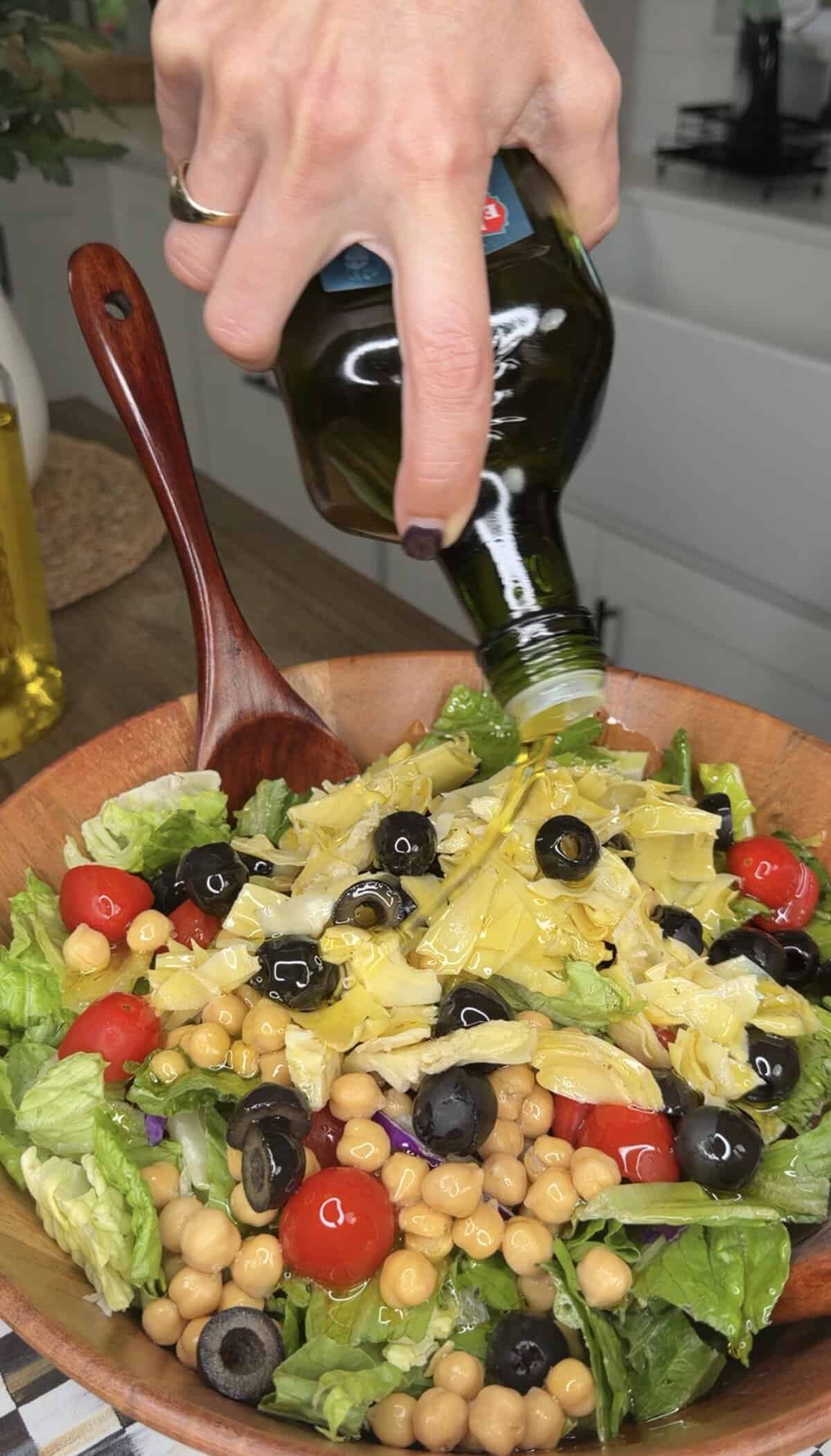 A hand pouring olive oil from a bottle onto a salad with lettuce, cherry tomatoes, chickpeas, black olives, artichoke hearts, and red onion in a wooden bowl alongside crispy Italian Fried Chicken.