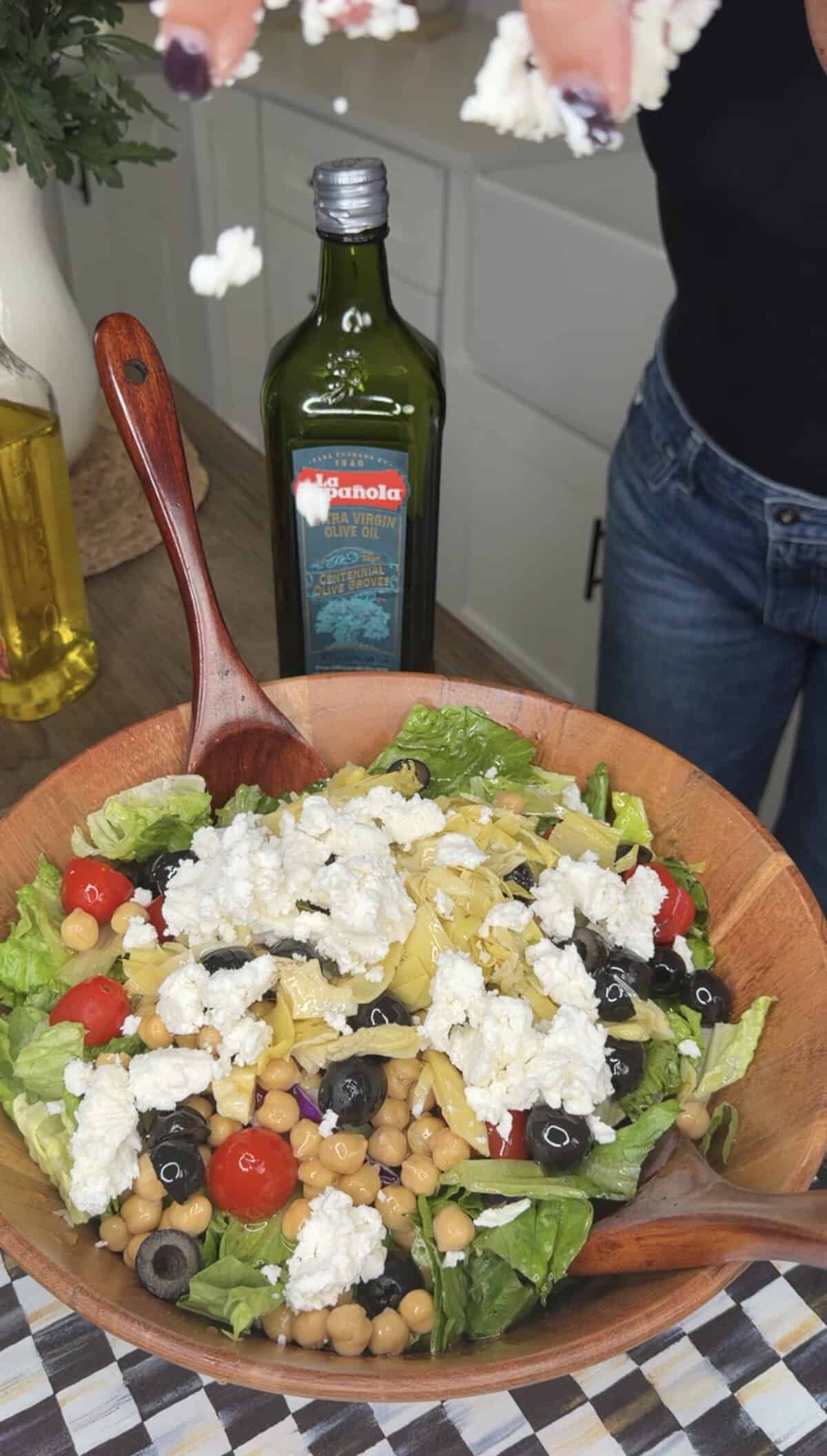 A wooden bowl filled with salad—lettuce, chickpeas, cherry tomatoes, black olives, artichoke hearts, and crumbled cheese—sits with a wooden spoon inside. Bottles of olive oil and canola oil hint at Italian Fried Chicken nearby.