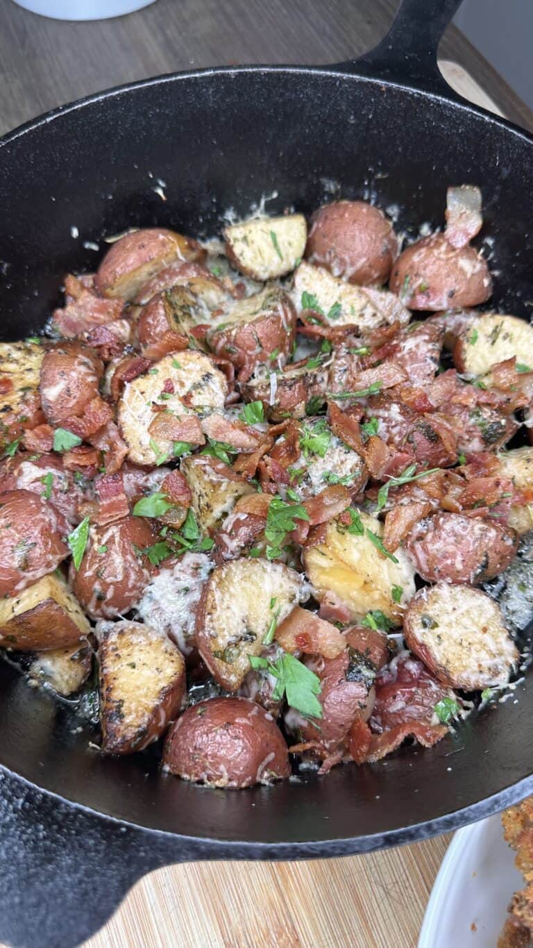 A cast iron skillet filled with Cast-Iron Potatos—roasted red potatoes topped with crispy bacon bits, melted cheese, and fresh chopped parsley. The skillet sits on a wooden surface.