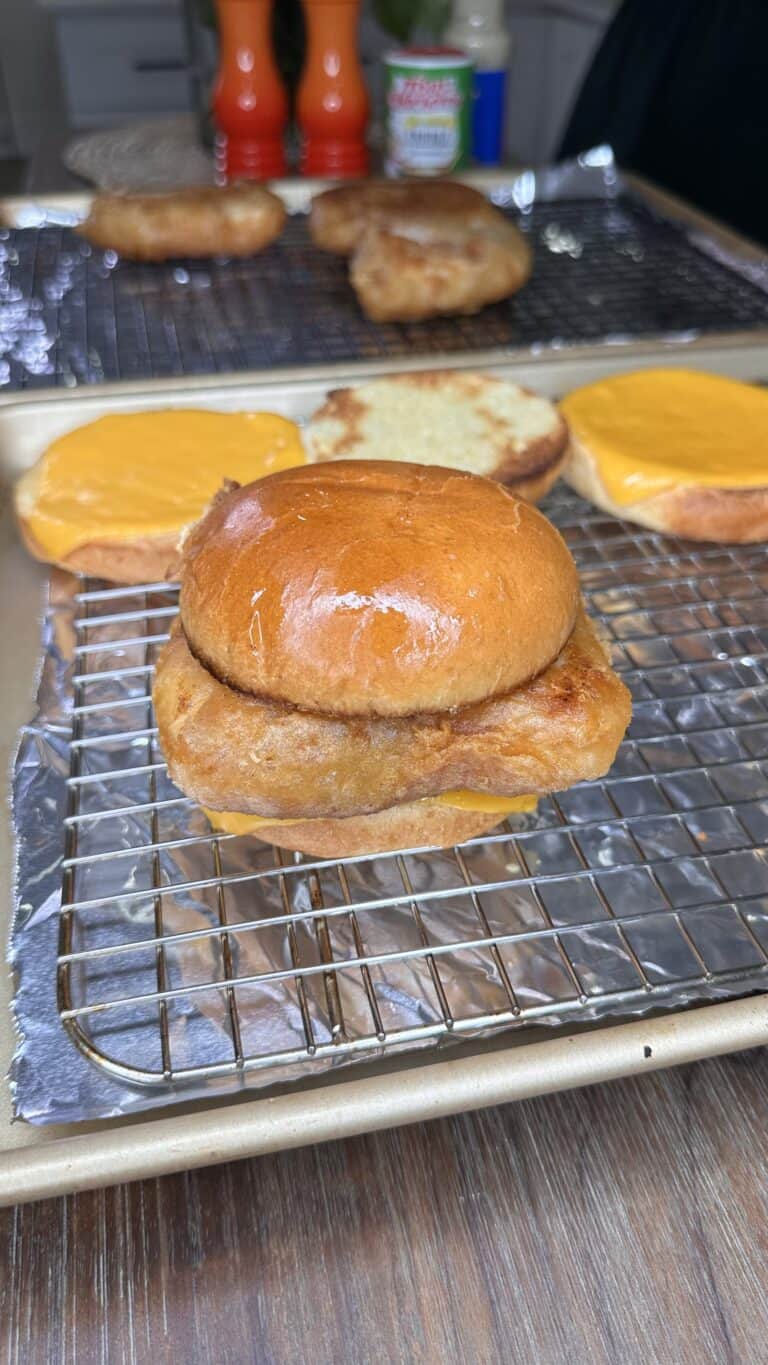 A Crispy Fish Filet Sandwich on a brioche bun sits on a wire rack, with sandwich buns and cheddar cheese slices in the background on a baking tray lined with foil.