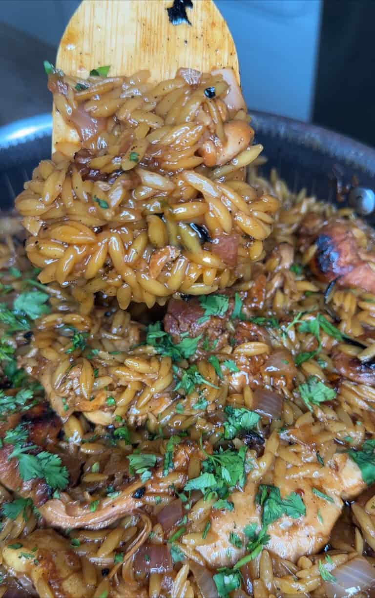 A close-up of a wooden spoon lifting a portion of cooked orzo mixed with Honey Lime Chicken, onions, herbs, and garnished with chopped fresh parsley in a pan.