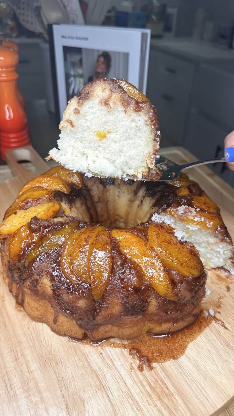 A Peach Upside Down Cake topped with caramelized peach slices sits on a wooden board. A slice is being lifted out, revealing a moist, light interior. The cake appears homemade and served in a cozy kitchen setting.