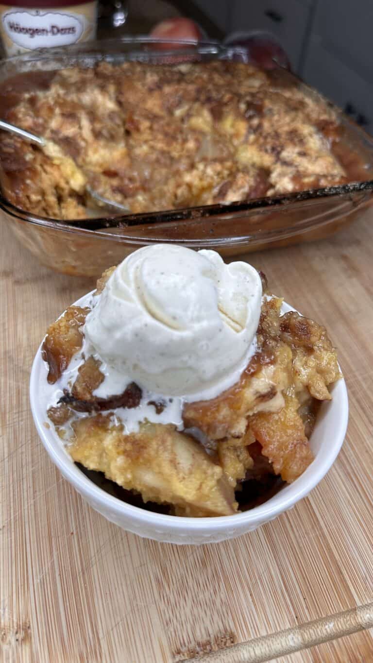 A bowl of Easy Peach Cobbler topped with a scoop of vanilla ice cream sits on a wooden surface, with a baking dish of more peach cobbler in the background.