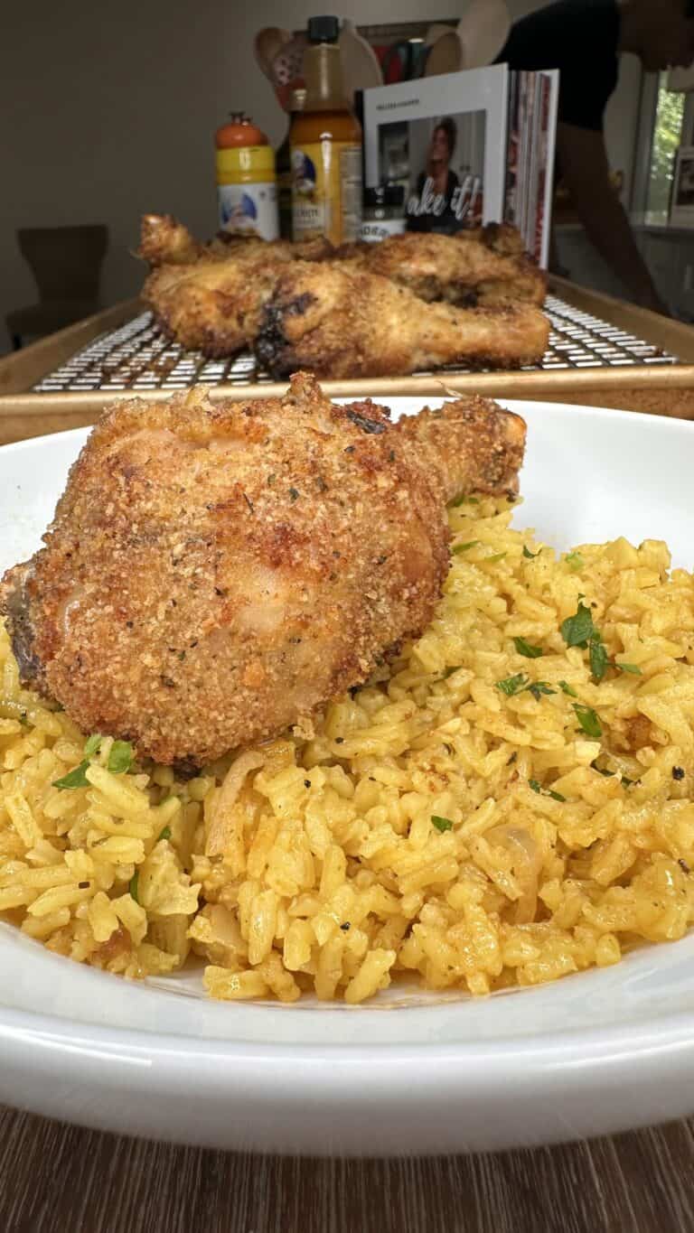 A crispy, golden-brown baked chicken drumstick sits atop a bed of vibrant Lemon Pepper Rice, garnished with herbs, in a white bowl. In the background, more chicken rests on a cooling rack.