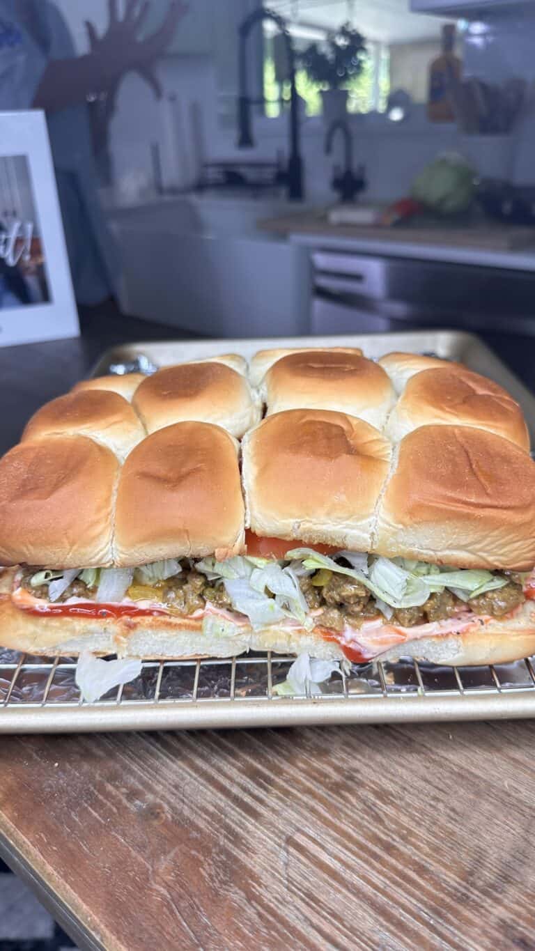 A tray of sliced Chopped Cheese slider sandwiches with golden brown buns, filled with lettuce, meat, and condiments, sits on a wooden table in a kitchen setting.