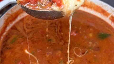 A ladle lifts a serving of cheesy, hearty Cheesy Stuffed Pepper Soup with visible chunks of tomato, beef, and spinach from a pot, with stretchy melted cheese trailing from the ladle back into the soup.