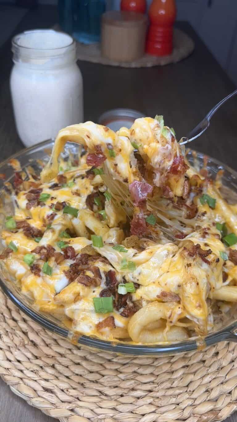A glass dish of Outback Cheese Fries & Ranch Copycat🍟 loaded with melted cheese, bacon bits, and green onions. A fork lifts a cheesy bite while a jar of creamy white sauce sits in the background.