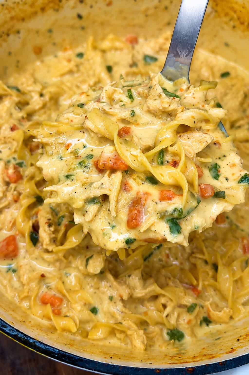 A close-up of a Creamy Chicken Noodle Soup 🍲 inspired pasta dish with visible pieces of chicken, pasta, chopped vegetables, and herbs, being scooped up with a large metal spoon.