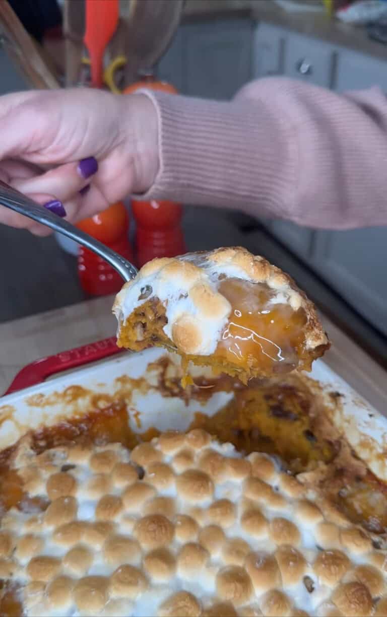 A hand with purple nail polish holds a spoonful of Sweet Potato Casserole 🍠 topped with toasted marshmallows over a baking dish of the same casserole in a cozy kitchen setting.