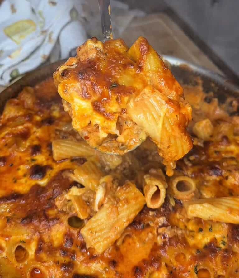 A close-up of One-Pot Million Dollar Rigatoni Bake with rigatoni noodles in a cheesy, browned tomato sauce. A spoon lifts a portion, revealing gooey melted cheese and the rich, saucy texture of this comforting dish.