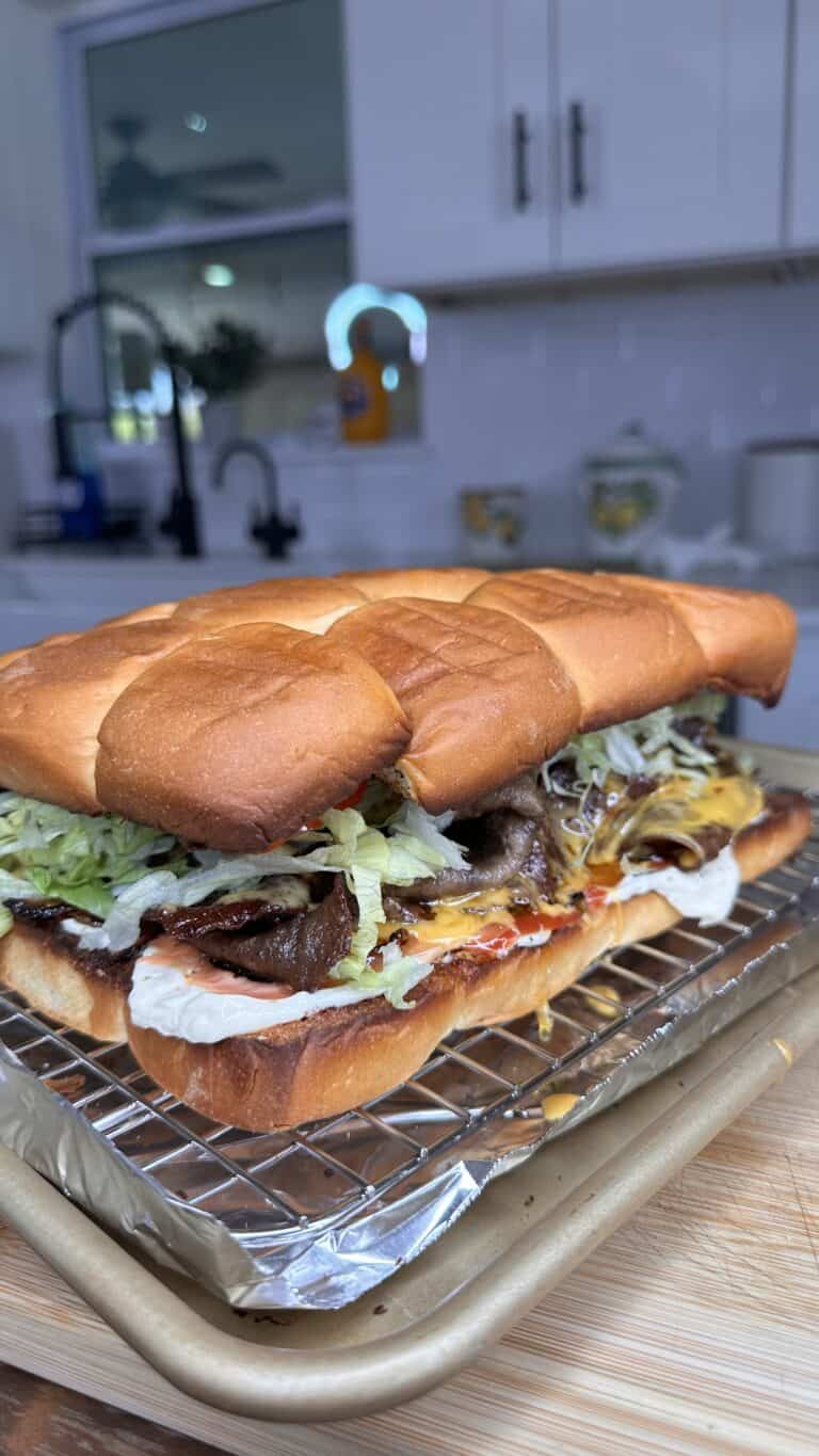 A toasted sub sandwich filled with sliced Steak Umm Sliders, melted cheese, shredded lettuce, and condiments sits on a wire rack over a baking sheet in a kitchen setting.