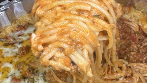 A close-up of a hand using a spatula to lift a cheesy, Million Dollar Spaghetti portion from a foil tray filled with baked spaghetti topped with melted cheese and tomato sauce.
