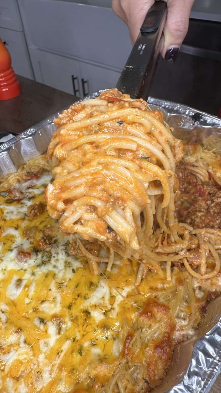 A close-up of a hand using a spatula to lift a cheesy, Million Dollar Spaghetti portion from a foil tray filled with baked spaghetti topped with melted cheese and tomato sauce.