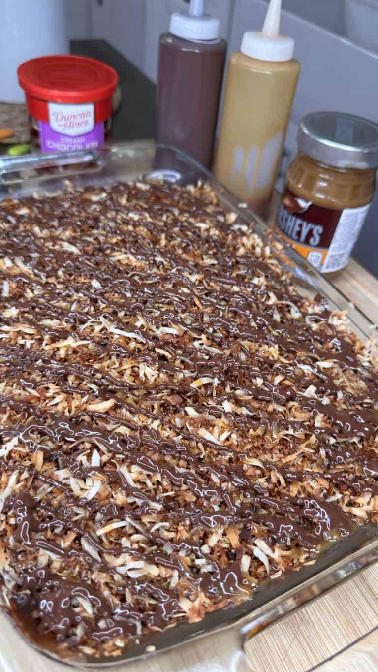 A glass baking dish filled with a dessert topped with toasted coconut, chocolate drizzle, and caramel sauce. Surrounding it are jars and bottles of chocolate syrup, caramel, and a tub of frosting.