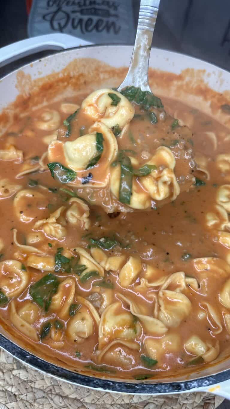 A ladle lifts cheese tortellini in Creamy Tomato Tortellini Soup with spinach from a pot, revealing a rich, thick broth and pasta generously coated in velvety tomato sauce.