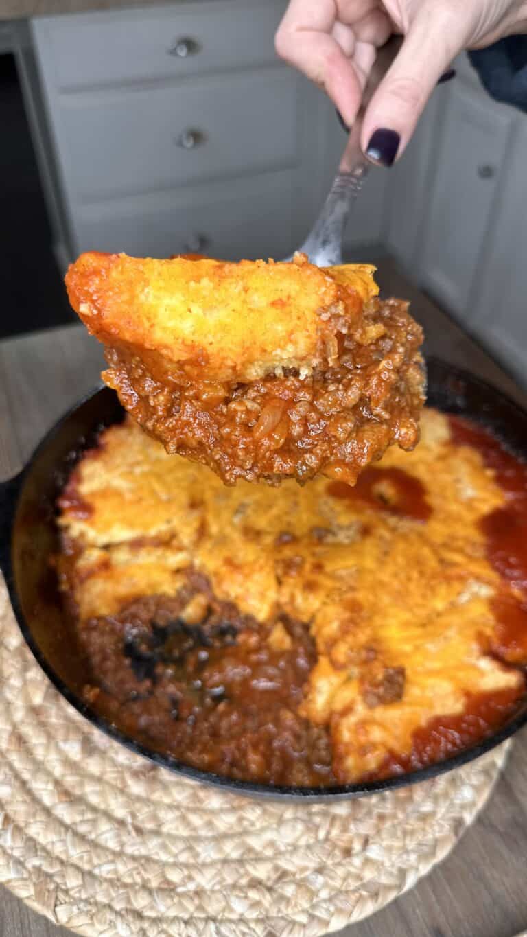 A hand holds a serving spoon with a portion of cheesy, baked Jiffy Joe chili cornbread casserole above a cast iron skillet filled with the same dish. The casserole has a golden, cheesy crust and rich, meaty filling.