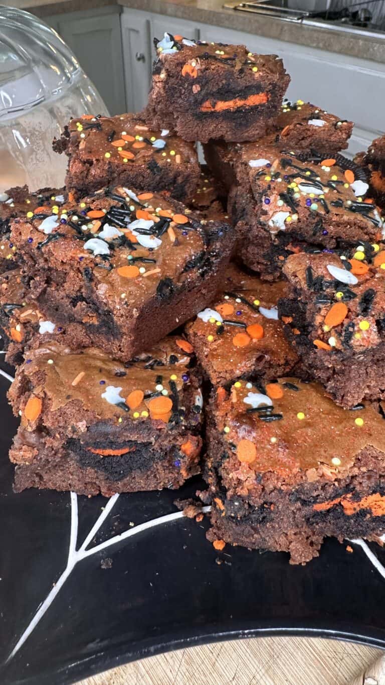 A stack of Halloween Slutty Brownies with Oreo cookies inside, topped with orange, white, black, and yellow Halloween sprinkles, arranged on a black platter with a white spiderweb design.