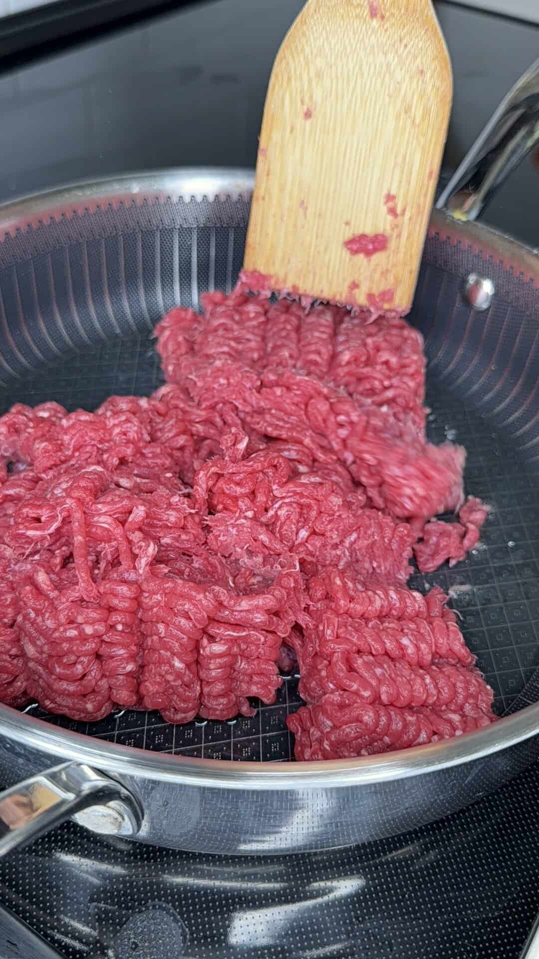 Raw ground beef is being broken up with a wooden spatula in a nonstick frying pan on a stovetop, the perfect first step for preparing a Beefy Creamy Taco Dip.