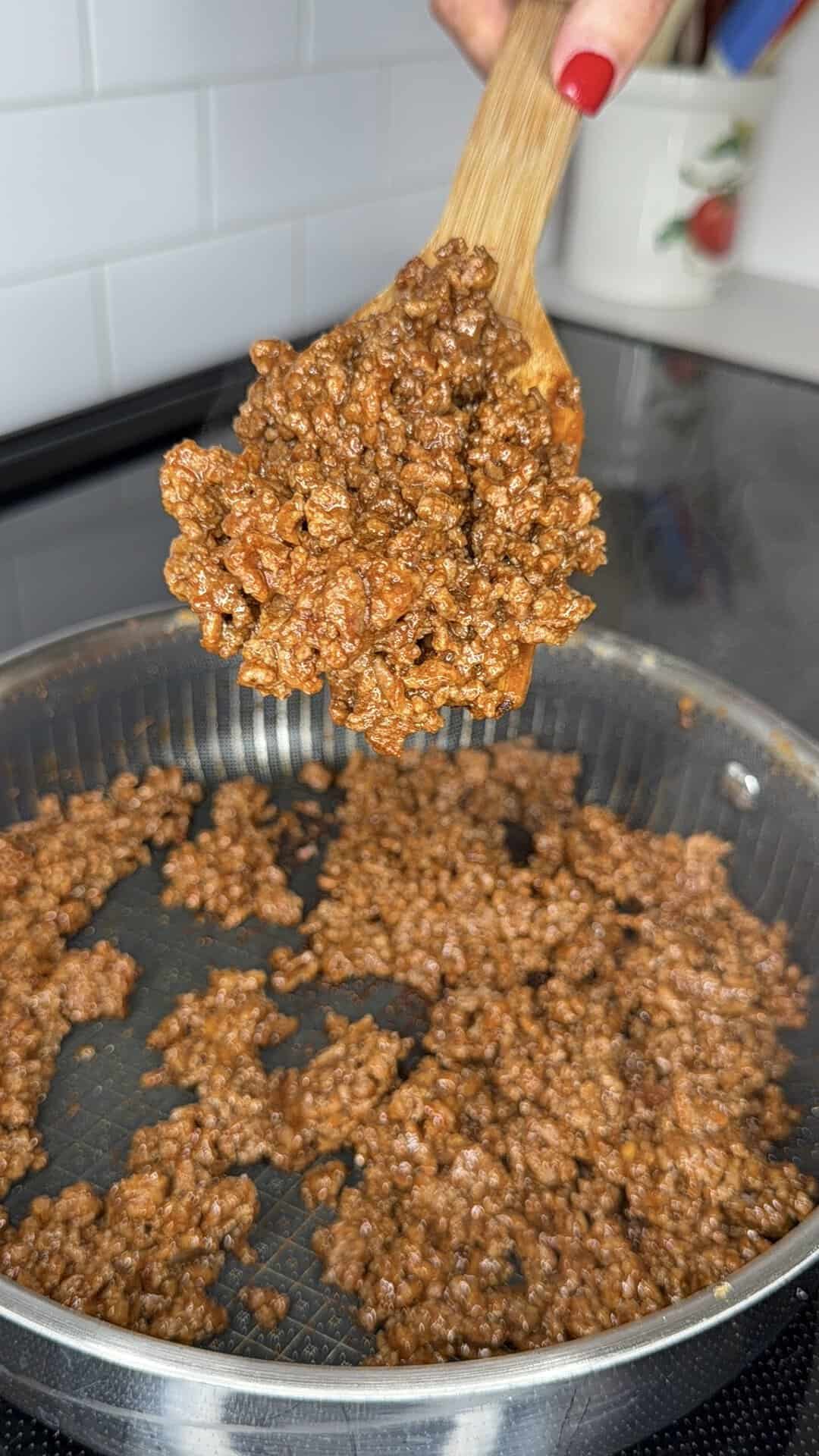 A hand holds a wooden spoon with cooked, seasoned ground beef for Beefy Creamy Taco Dip over a pan filled with more of the same beef on a stovetop.