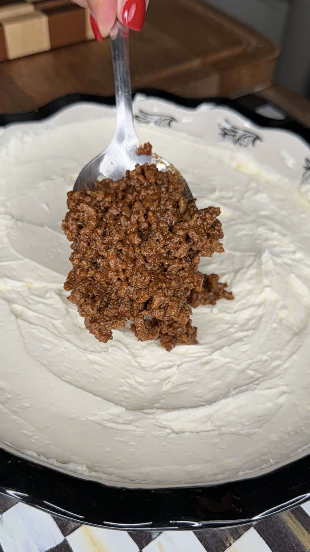 A hand holds a spoon with seasoned ground beef over a dish spread with cream cheese, preparing to add the savory meat layer for a delicious Beefy Creamy Taco Dip.