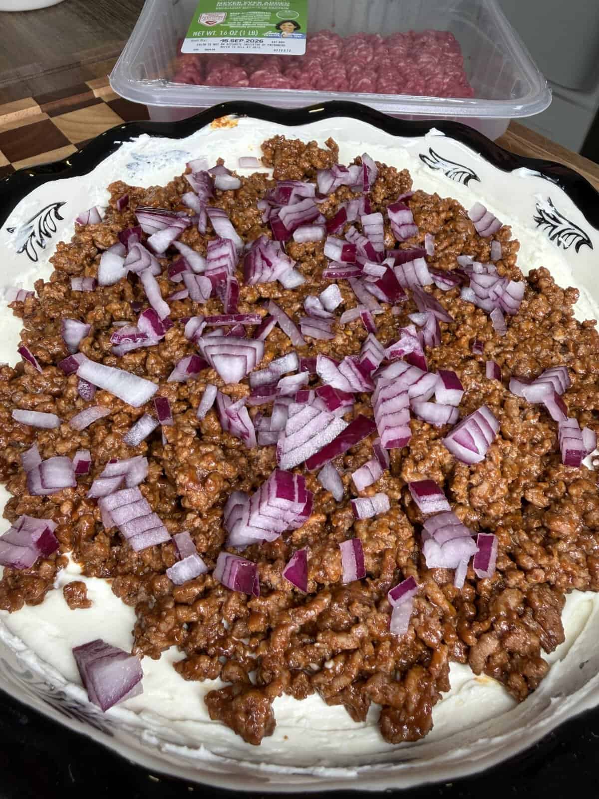 A close-up of Beefy Creamy Taco Dip features a creamy white dip topped with seasoned ground beef and chopped red onions, with a package of raw ground beef in the background.