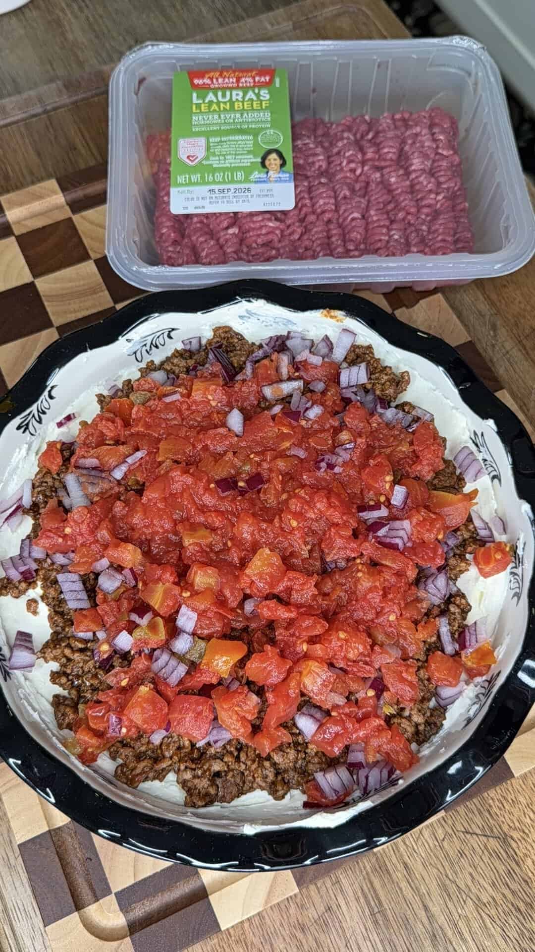 A pie dish with ground beef, chopped red onions, and diced tomatoes sits on a wooden surface, reminiscent of the flavors found in a Beefy Creamy Taco Dip. Nearby, there is a package of ground lamb and a cutting board.