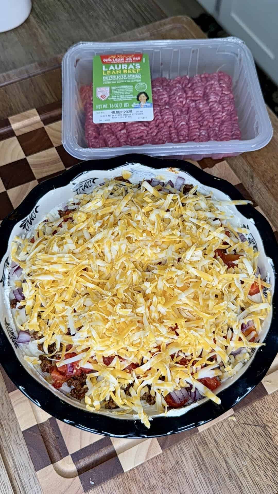 A pie dish filled with an unbaked casserole, soon to become a Beefy Creamy Taco Dip, is topped with shredded cheese and rests on a wooden cutting board. Behind it is a package of raw ground beef, ready to add hearty flavor to this delicious dish.