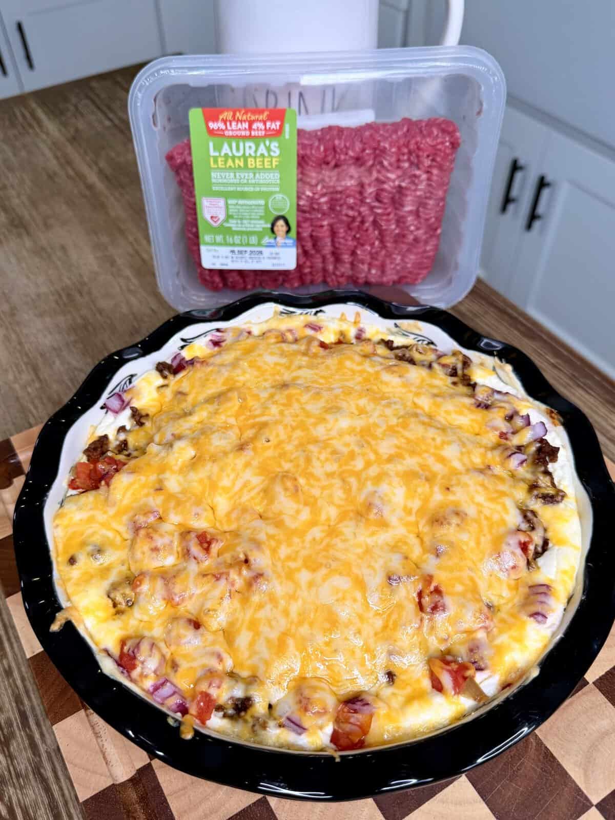 A baked casserole topped with melted cheese sits in front of a package of raw ground beef on a kitchen counter. Reminiscent of a hearty Beefy Creamy Taco Dip, the casserole appears to have beef, red onions, and tomatoes layered under the gooey cheese.