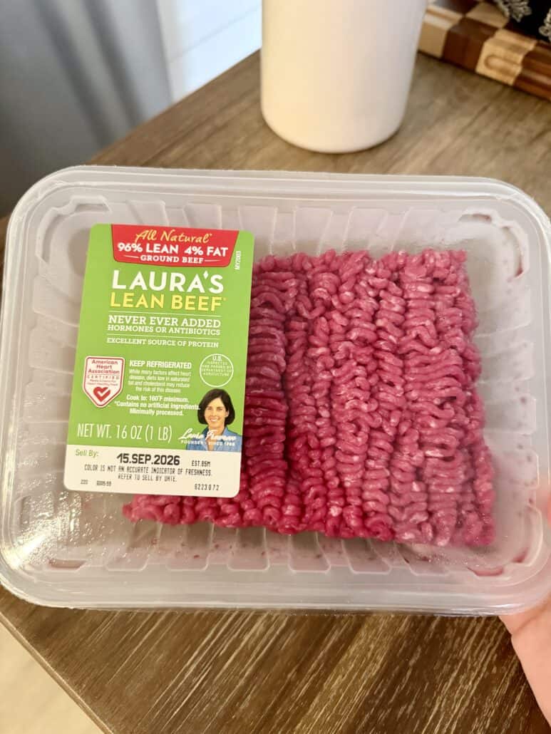 A hand holds a plastic package of Lauras Lean Beef ground beef, labeled 96% lean, 4% fat—perfect for recipes like Beefy Creamy Taco Dip. The package weighs 16 oz and has a green label with storage and expiration information.