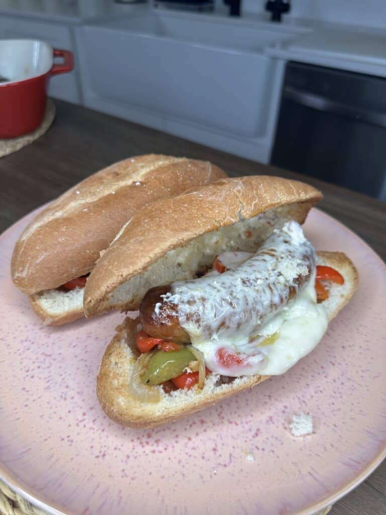 An Easy Bake Sausage and Peppers creation: a sausage in a bun topped with melted cheese, grilled peppers, and onions sits on a pink plate, with another split bun beside it on the kitchen counter.