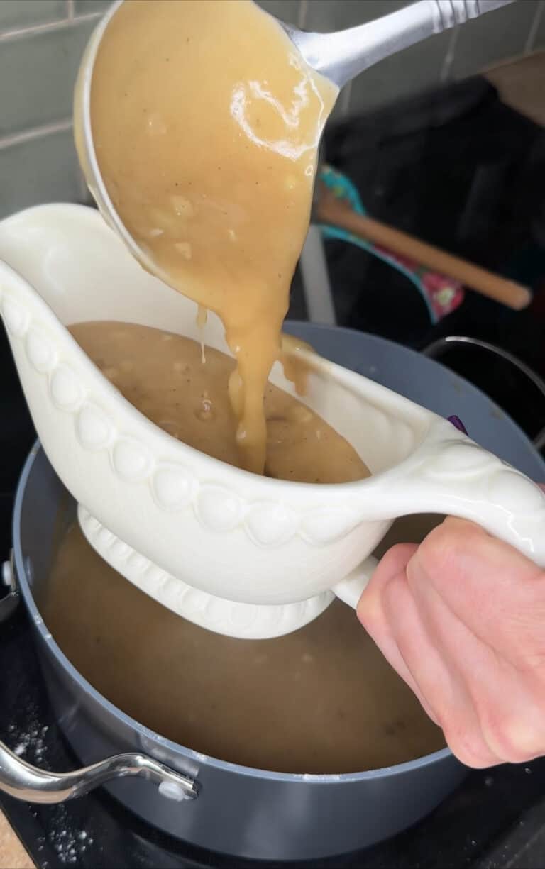 A hand holds a white gravy boat as thick, brown Turkey Gravy 🦃 is ladled from a saucepan on the stove, ready to serve.