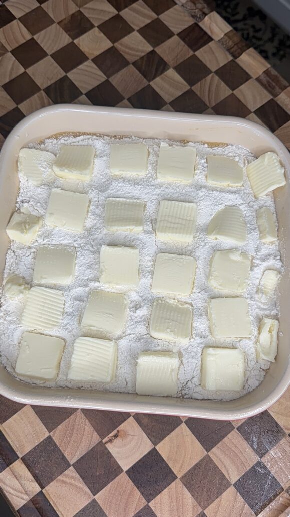 A baking dish with a layer of white flour topped with evenly spaced, thin slices of butter, ready to become the base for a delicious Pumpkin Pie Cake, sits on a checkered wooden surface.