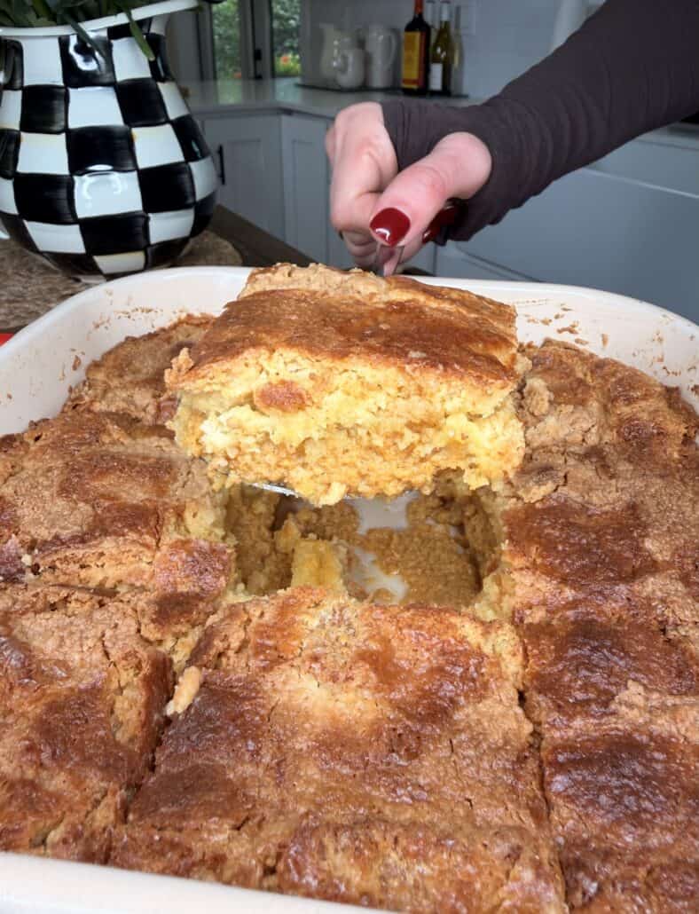 A hand cuts a square piece from a golden, crumbly Pumpkin Pie Cake in a white dish. A black-and-white checkered pitcher is in the background on a kitchen counter.