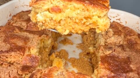 A hand with red nail polish lifts a serving of golden, crumbly cornbread from a baking dish, surrounded by cut pieces reminiscent of Pumpkin Pie Cake. A black and white checkered vase is in the background.