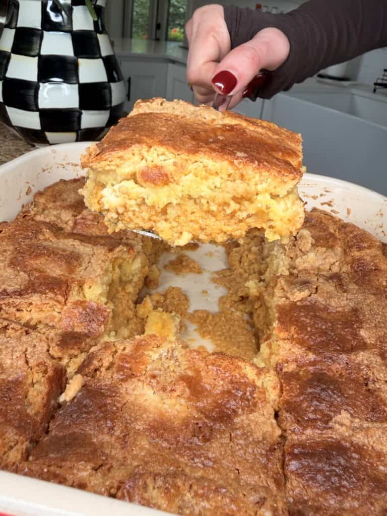 A hand with red nail polish lifts a serving of golden, crumbly cornbread from a baking dish, surrounded by cut pieces reminiscent of Pumpkin Pie Cake. A black and white checkered vase is in the background.